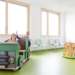 Zwei Kinder im grünen Spielauto im Wartebereich des KÄZ Dornbirn.
