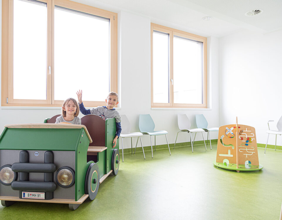 Zwei Kinder im grünen Spielauto im Wartebereich des KÄZ Dornbirn.